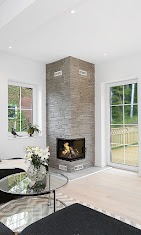 Wnętrze salonu z narożnym kominkiem obłożonym szarą cegłą, płomienie widoczne za szybą, jasne ściany i podłoga, okno z widokiem na zieleń, szklany stolik kawowy z bukietem kwiatów.