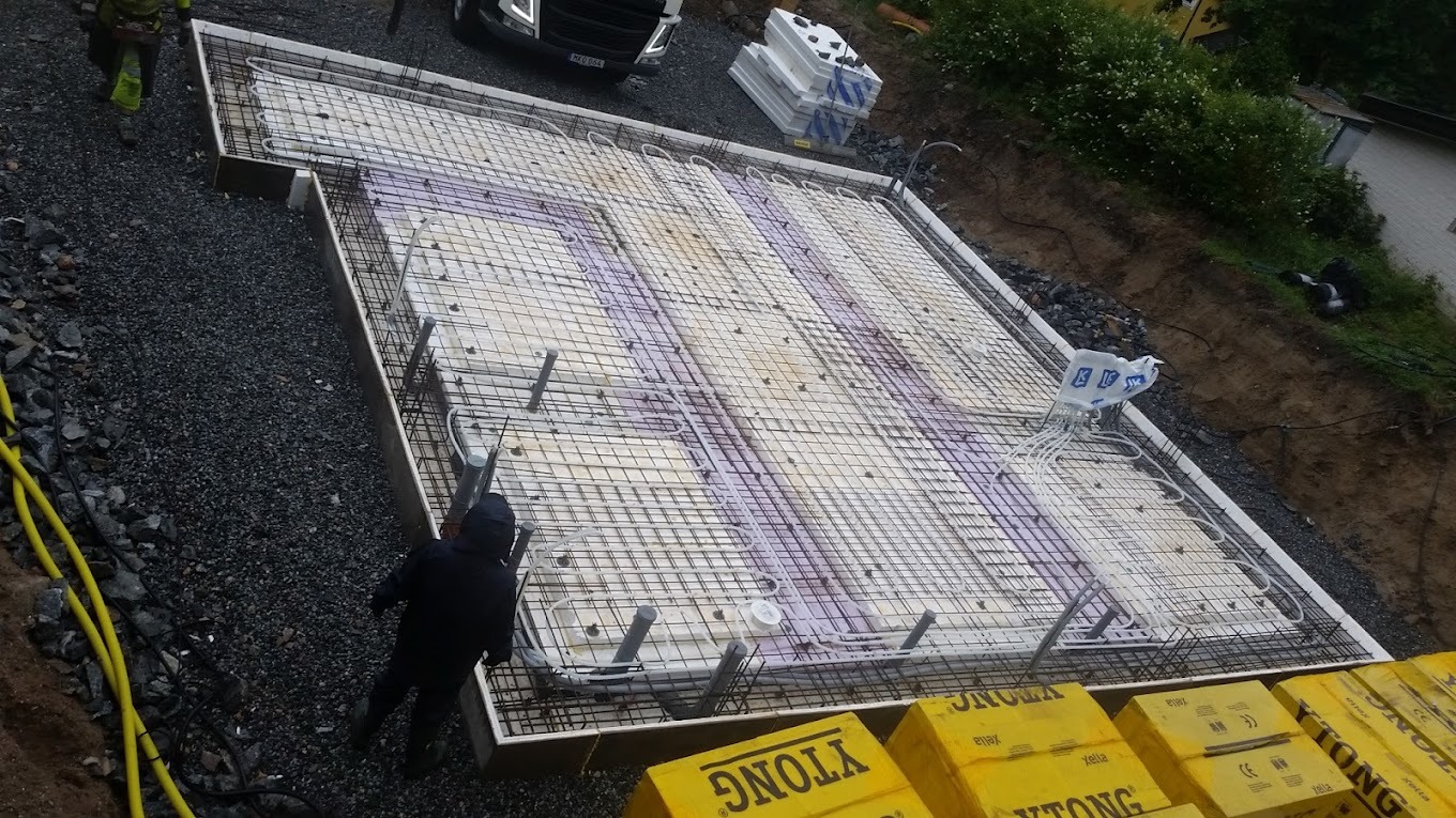 Przygotowanie płyty fundamentowej z instalacją ogrzewania podłogowego, widoczne rury grzewcze ułożone na warstwie izolacyjnej, zbrojenie z siatki stalowej, szalunek obwodowy i stos bloczków Ytong.
