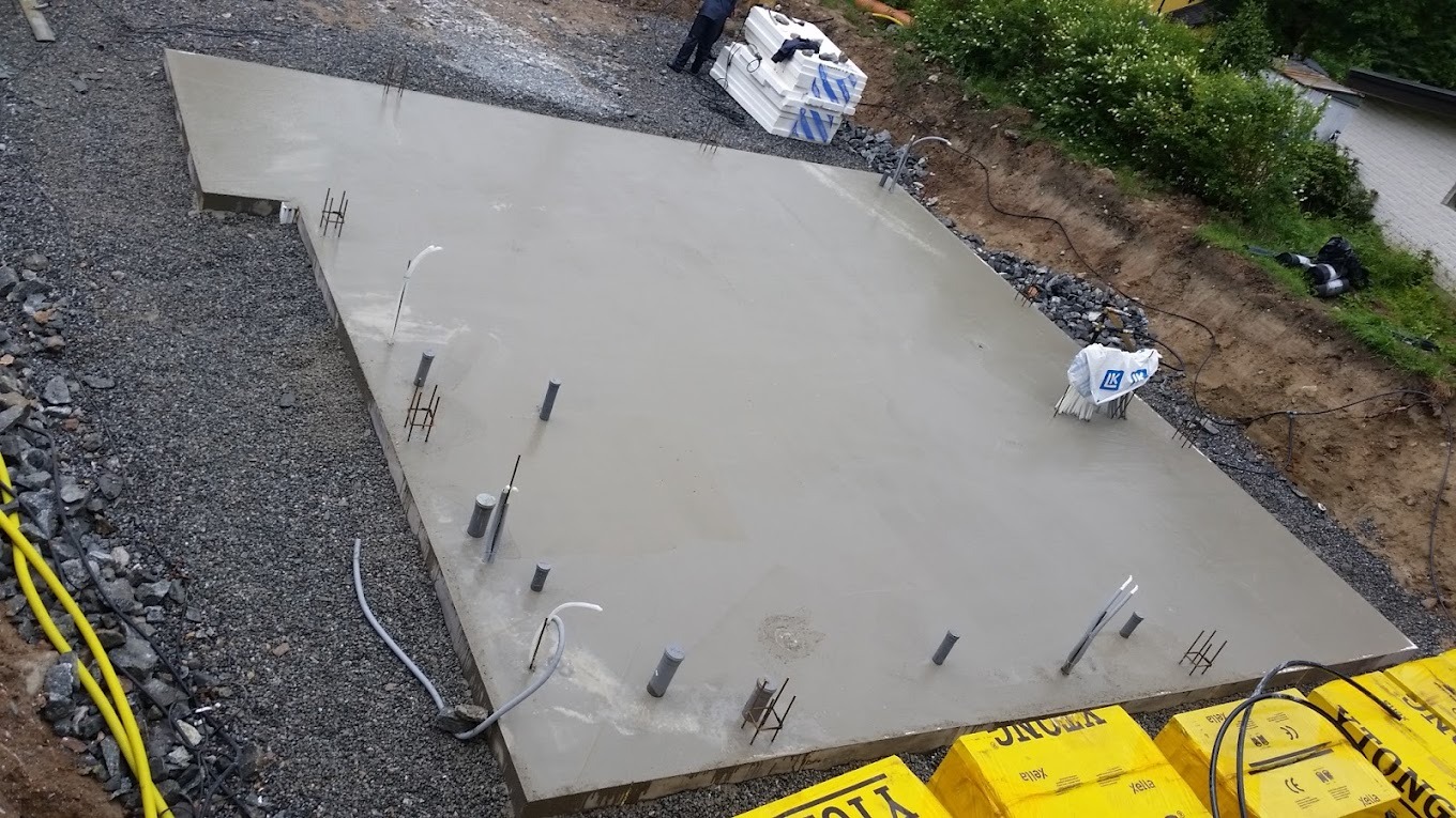 Świeżo wylana, gładka płyta fundamentowa z wystającymi rurami instalacyjnymi i prętami zbrojeniowymi, widok z góry na plac budowy z przygotowanym materiałem izolacyjnym.