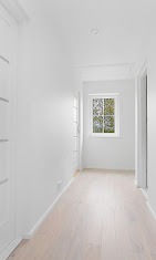 Jasny korytarz z białymi ścianami, panelami podłogowymi w odcieniach beżu, białymi drzwiami i oknem z widokiem na zieleń.