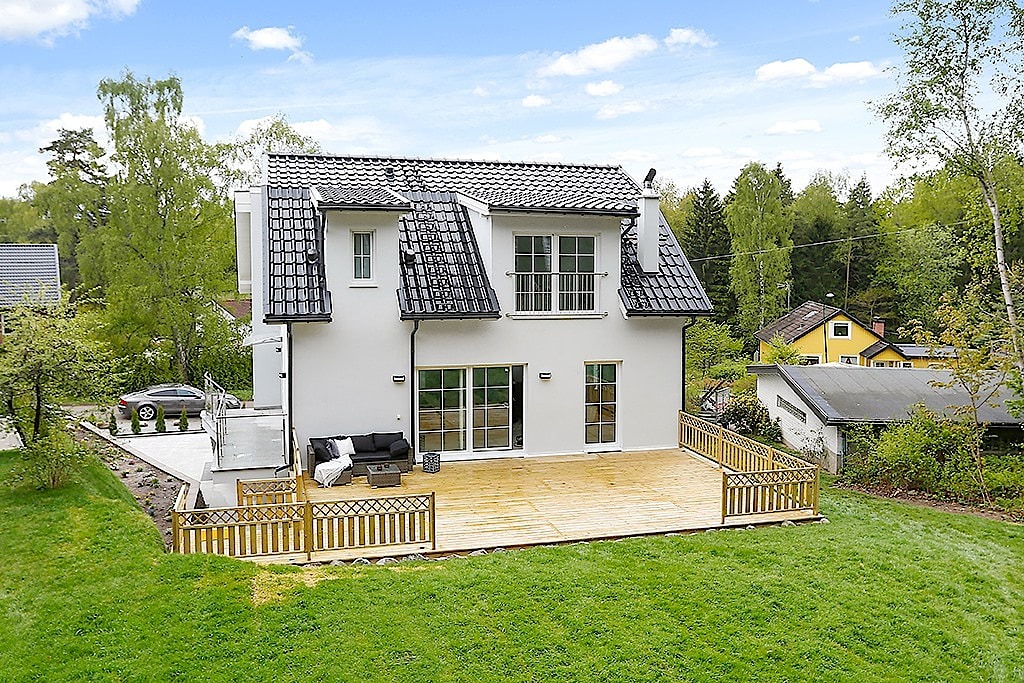 Nowoczesny dom z białymi ścianami, czarnym dachem z dachówki karpiówki i drewnianym tarasem z ażurową balustradą, widok z ogrodu w słoneczny dzień.