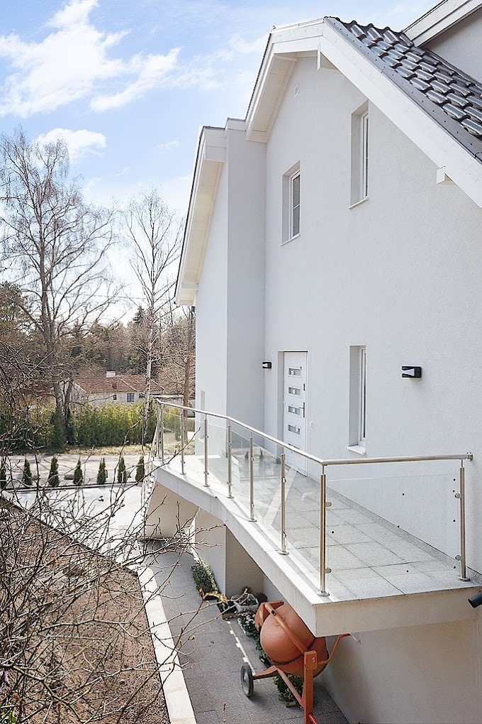 Biały dom z balkonem z przeszkloną balustradą, widoczna również pomarańczowa betoniarka pod balkonem i fragment dachu z szarej dachówki.