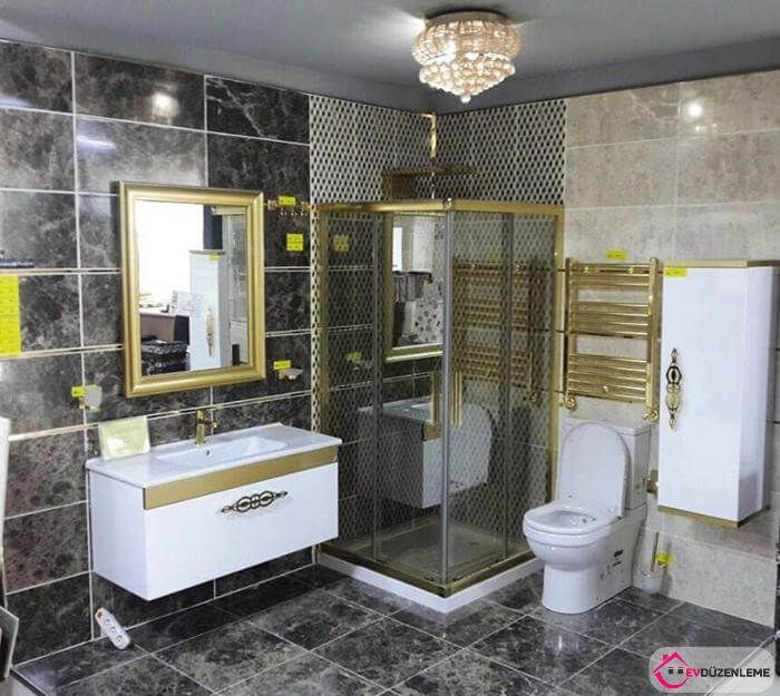 Nowoczesna łazienka z marmurowymi płytkami, złotymi akcentami na armaturze, kabinie prysznicowej i grzejniku, oraz kryształowym żyrandolem.