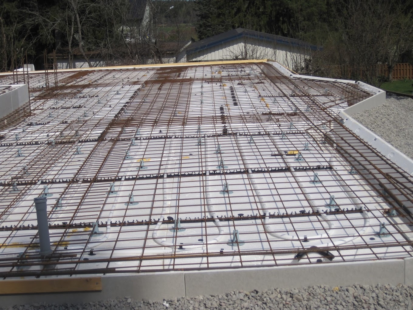 Przygotowanie pod wylewkę fundamentu: siatka zbrojeniowa na styropianie z wbudowanymi rurami ogrzewania podłogowego, widoczne szare rury kanalizacyjne i obrzeże z betonu.