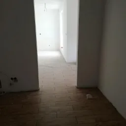 Ułożona podłoga z płytek imitujących drewno w pustym, odnowionym pokoju z surowymi ścianami i widocznymi instalacjami elektrycznymi.