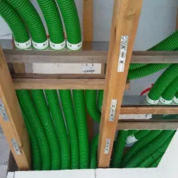 Zielone, karbowane rury instalacyjne przymocowane do drewnianej konstrukcji dachu za pomocą metalowych uchwytów, widoczne białe bloki konstrukcyjne.