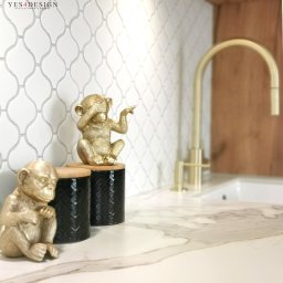 YES4DESIGN Elwira Bowycz-Stryjek - Biała mozaika w kształcie łezki na ścianie nad blatem z marmurowym wzorem; złota bateria umywalkowa i dekoracyjne figurki małp w złotym kolorze na czarnych pojemnikach z bambusowymi pokrywkami.