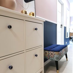 YES4DESIGN Elwira Bowycz-Stryjek - Szary kredens na buty z dekoracyjnymi, granatowymi gałkami, obok tapicerowana pufa z geometrycznym wzorem, na ścianie róż i granat, w tle drzwi i dekoracje.