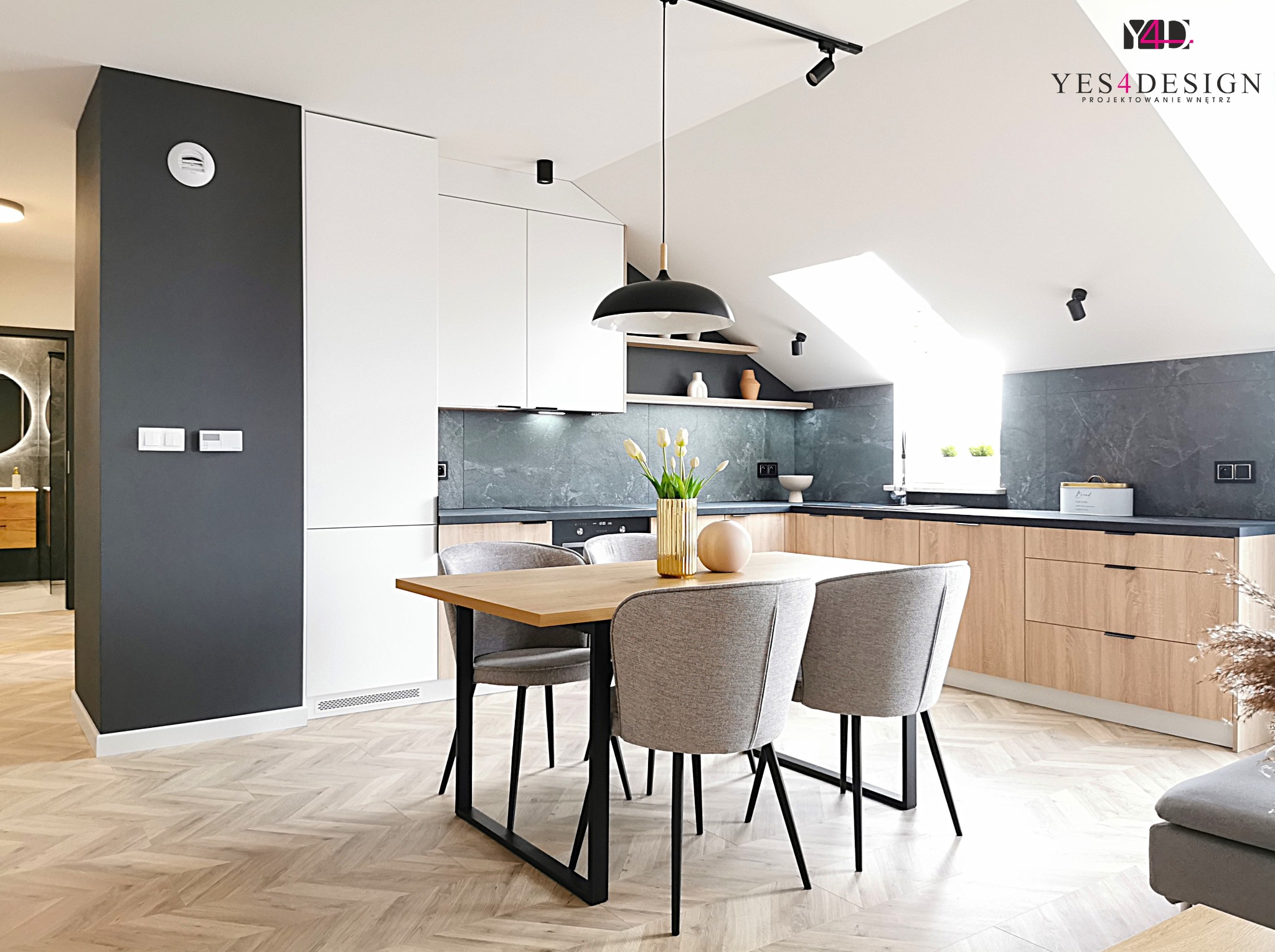 Jasna kuchnia z drewnianymi szafkami, szarym blatem i marmurową ścianą, stół z szarymi krzesłami, parkiet w jodełkę, czarna lampa wisząca i reflektory szynowe.