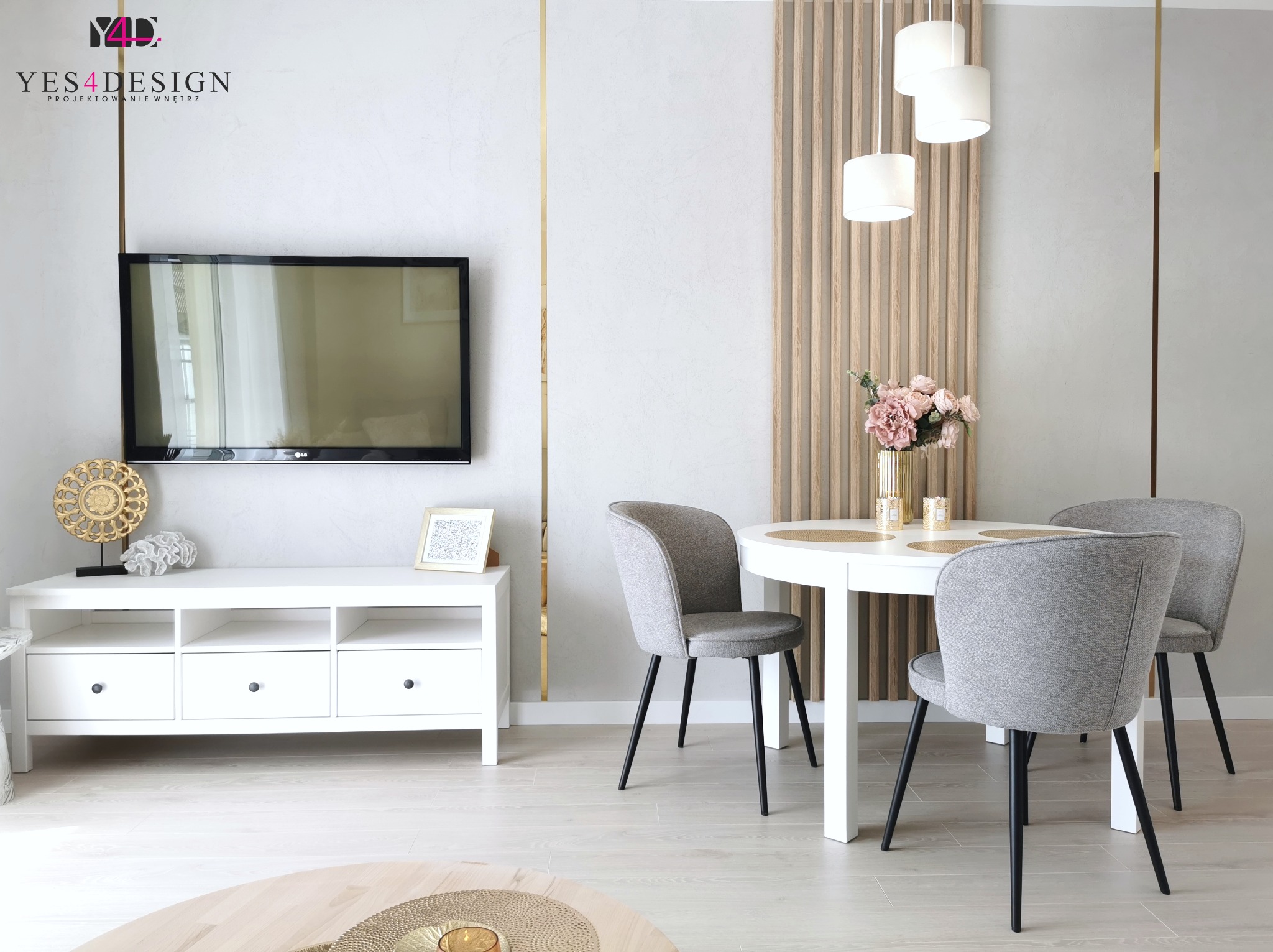 Jasne wnętrze jadalni z białym okrągłym stołem, szarymi krzesłami na czarnych nogach, drewnianą ścianką działową, telewizorem na ścianie i białymi meblami pod nim, oświetlone trzema lampami wiszącymi.