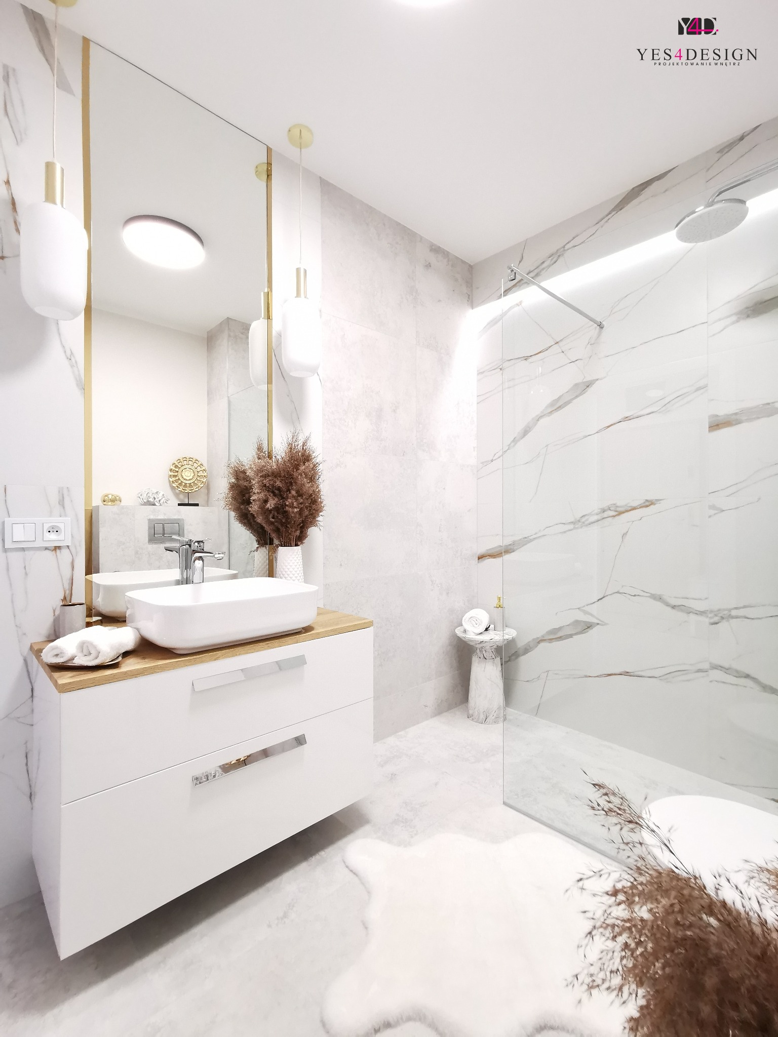 Jasna łazienka z marmurowymi płytkami, białą umywalką nablatową na drewnianym blacie i kabiną prysznicową ze szklaną ścianką, oświetlona złotymi lampami wiszącymi.