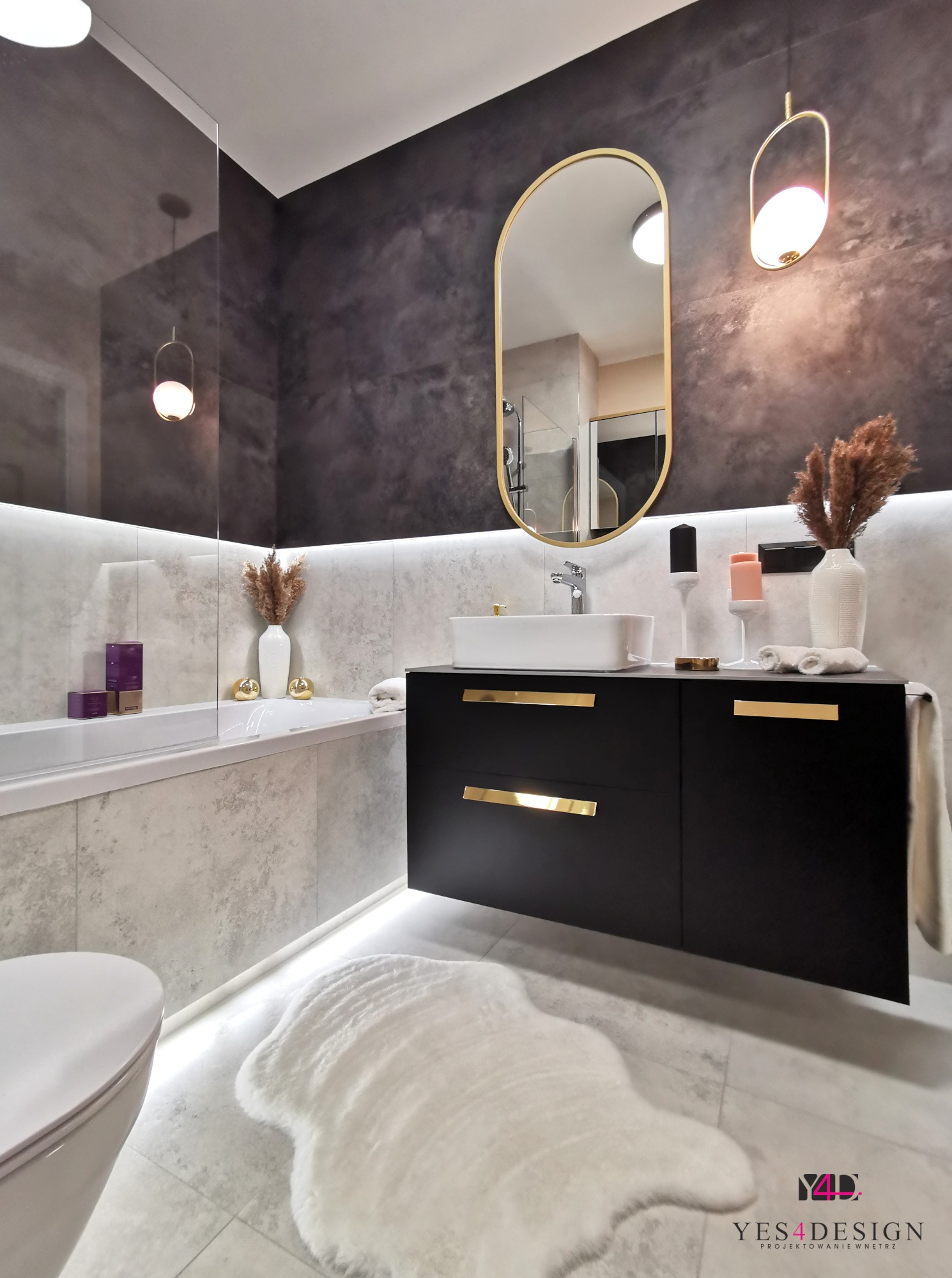Nowoczesna łazienka z czarną szafką pod umywalką, złotymi uchwytami, lustrem w złotej ramie, oświetleniem LED pod wanną i dekoracjami, w tym biały wazon z suszonymi trawami.
