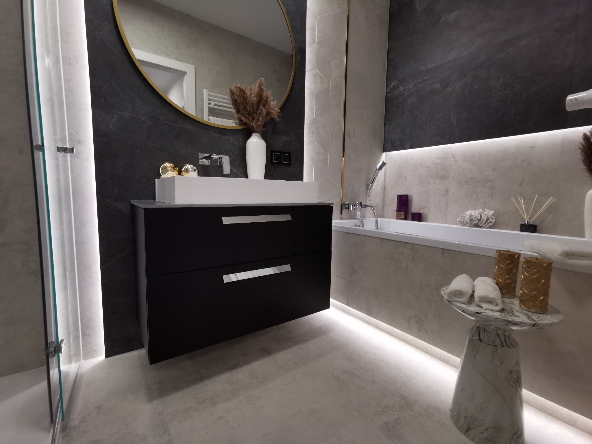Nowoczesna łazienka z czarną szafką pod umywalką, owalnym lustrem w złotej ramie, marmurową ścianą i podświetlaną wanną.