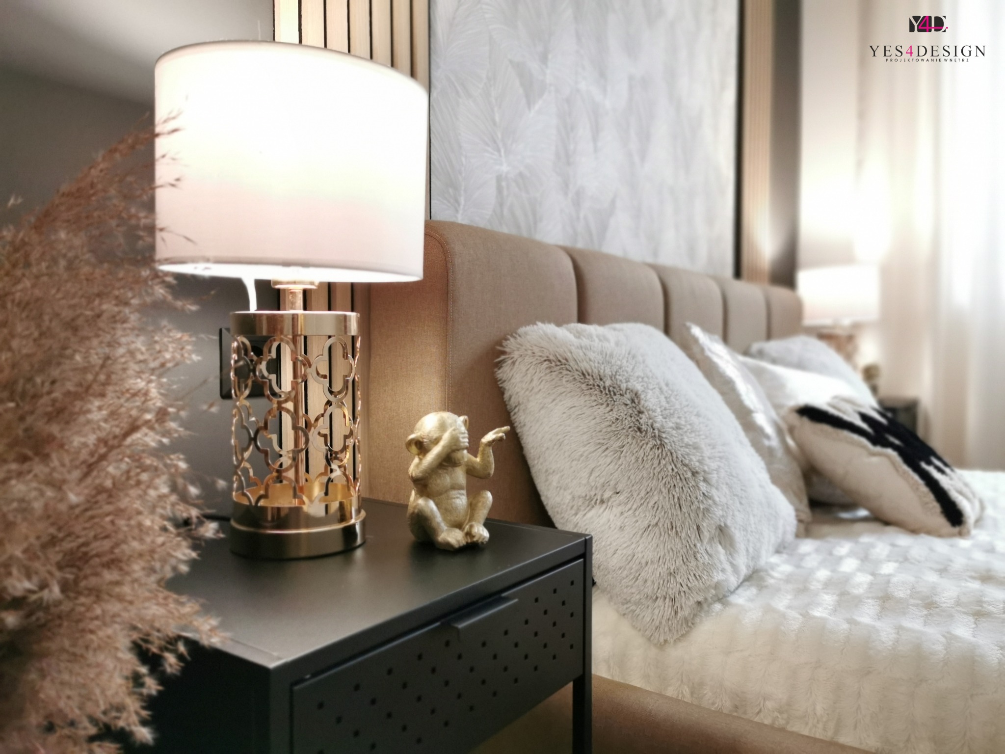 Sypialnia z łóżkiem z tapicerowanym zagłówkiem, lampką nocną z ażurową podstawą, figurką małpki i puszystymi poduszkami, dekorowana trawą pampasową i tapetą w liście.