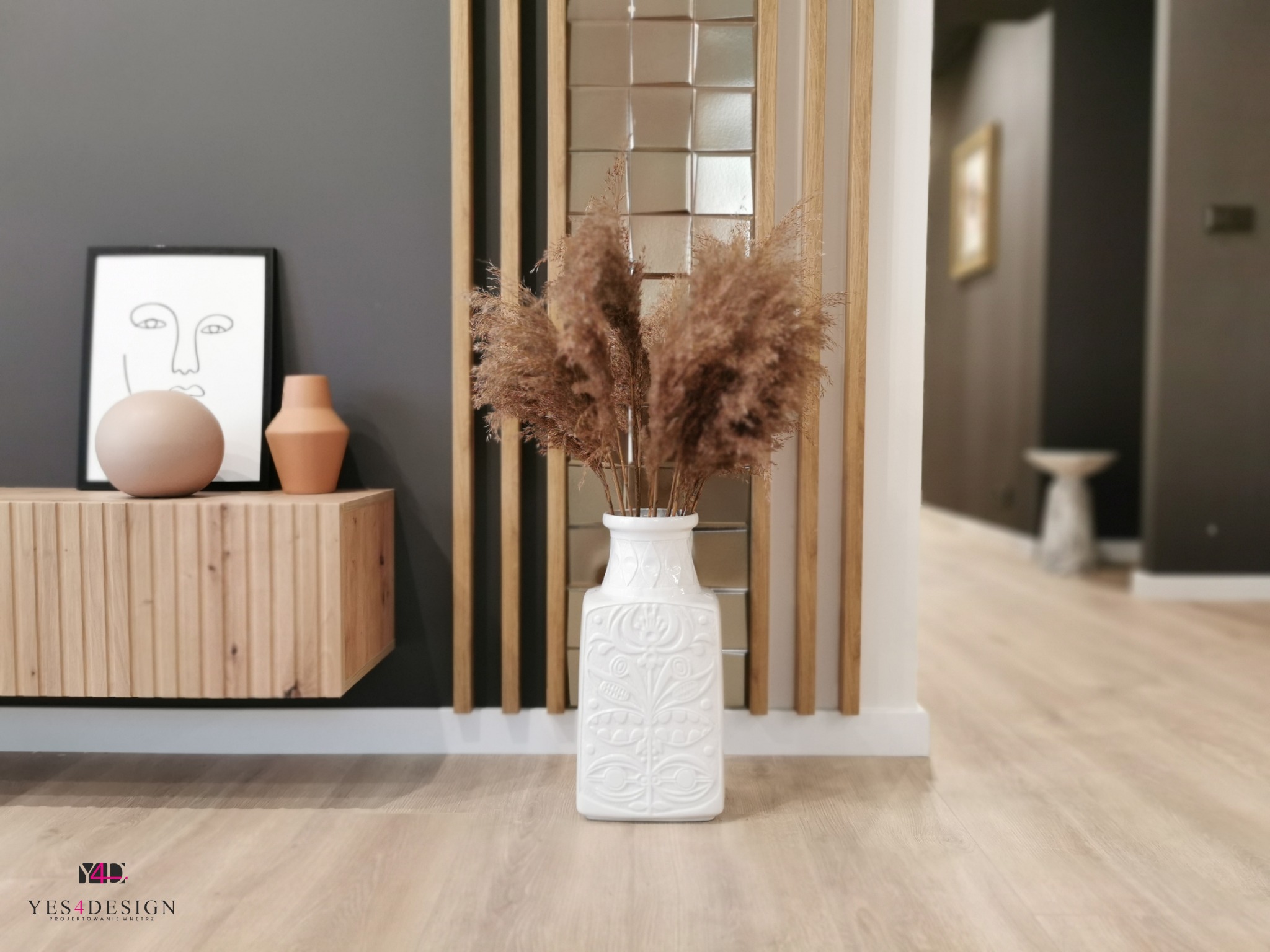Nowoczesne wnętrze: drewniana szafka RTV z dekoracjami (abstrakcyjny portret, ceramiczne kule i wazoniki) pod szarą ścianą, biały wazon z suszoną trawą na parkiecie, drewniana ażurowa ścianka...