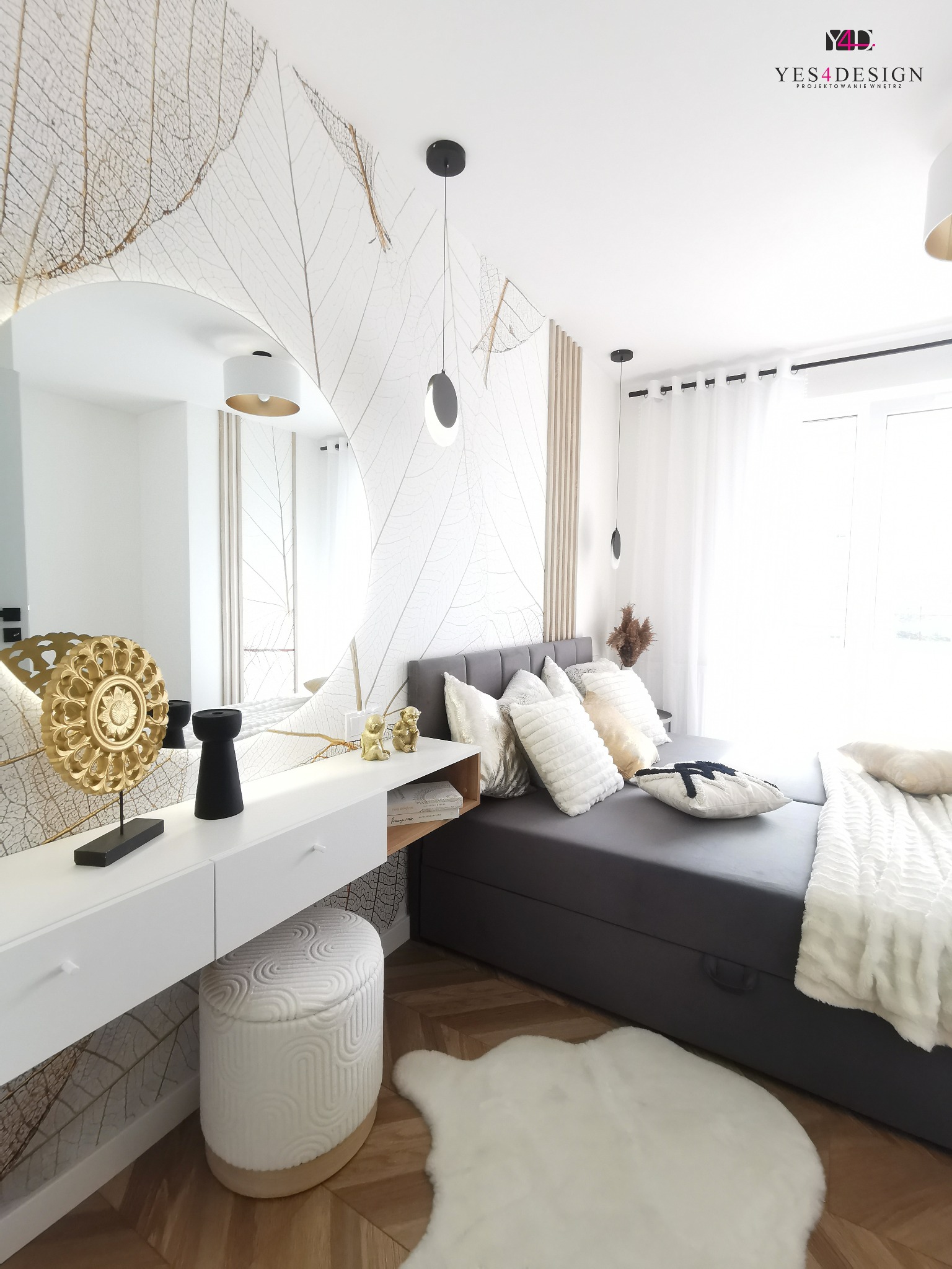 Jasna sypialnia z łóżkiem kontynentalnym, toaletką z okrągłym lustrem i tapetą w motywy liści; na toaletce złota dekoracja i figurki, na podłodze dywanik z długiego włosia.