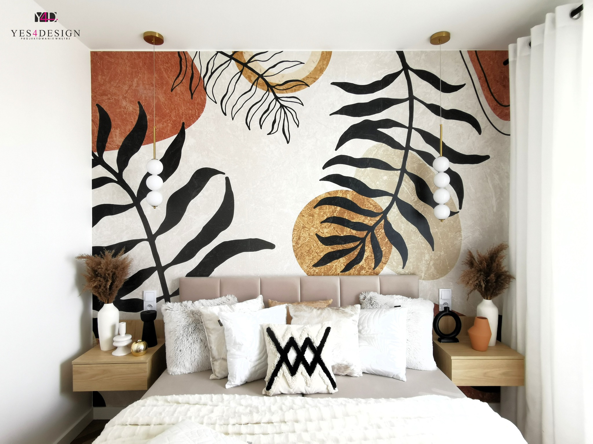 Sypialnia z łóżkiem, jasną pościelą i poduszkami dekoracyjnymi, na ścianie abstrakcyjny wzór z motywami roślinnymi w kolorach beżu, brązu i czerni, wiszące lampy z białymi kulami po bokach łóżka...