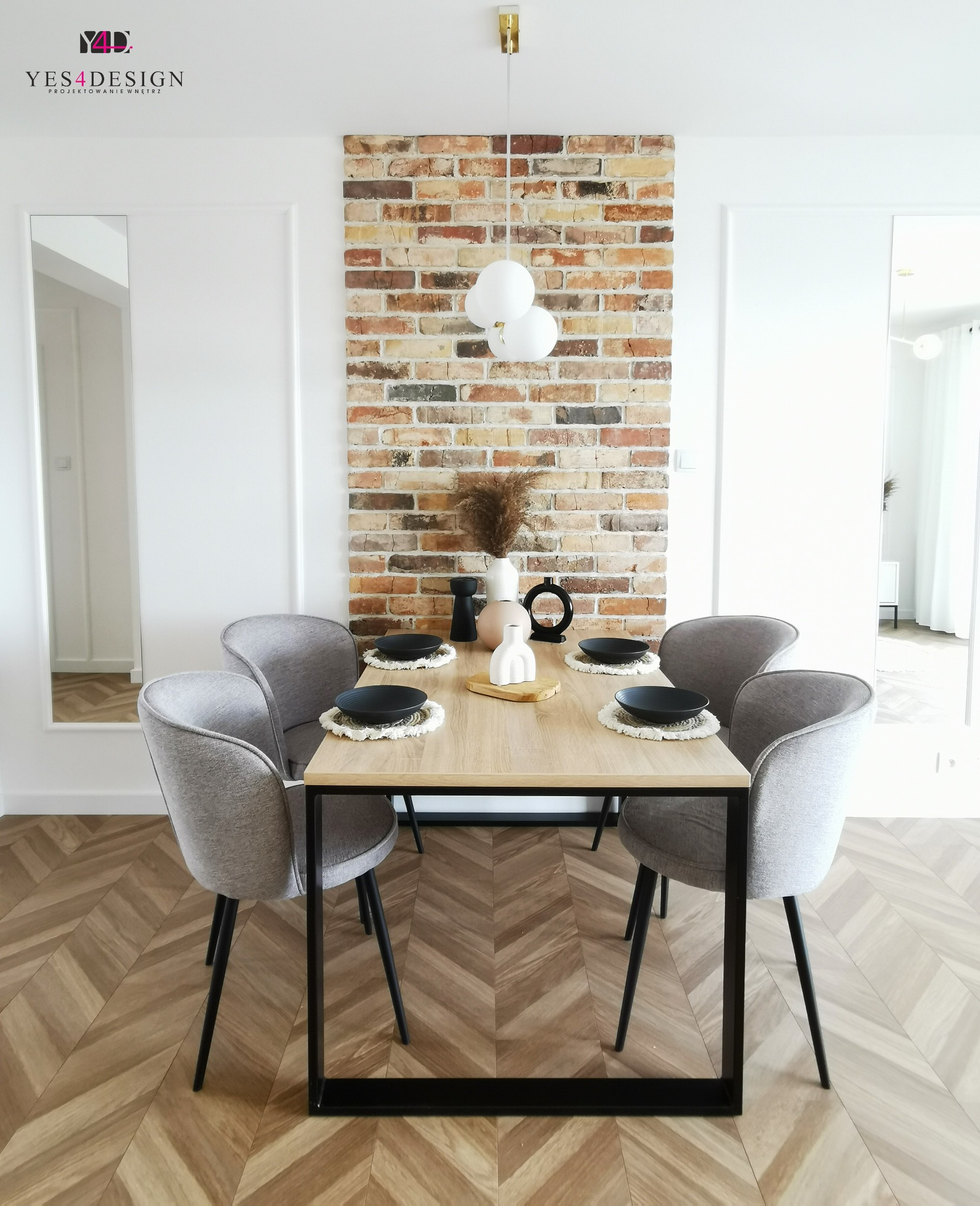 Jadalnia w stylu minimalistycznym z drewnianym stołem na czarnych nogach, szarymi krzesłami, dekoracyjną ścianą z cegły i podłogą w jodełkę, oświetlona trzema białymi kulami.