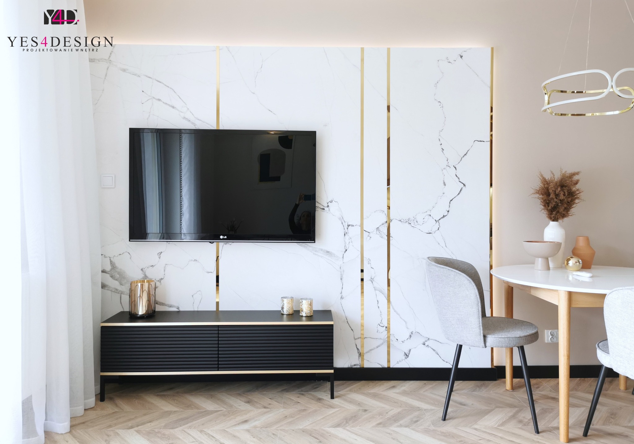 Salon z telewizorem na ścianie z marmurowym wzorem i złotymi wstawkami, czarna szafka RTV z dekoracjami, okrągły stół z szarymi krzesłami i suszonymi kwiatami w wazonie.