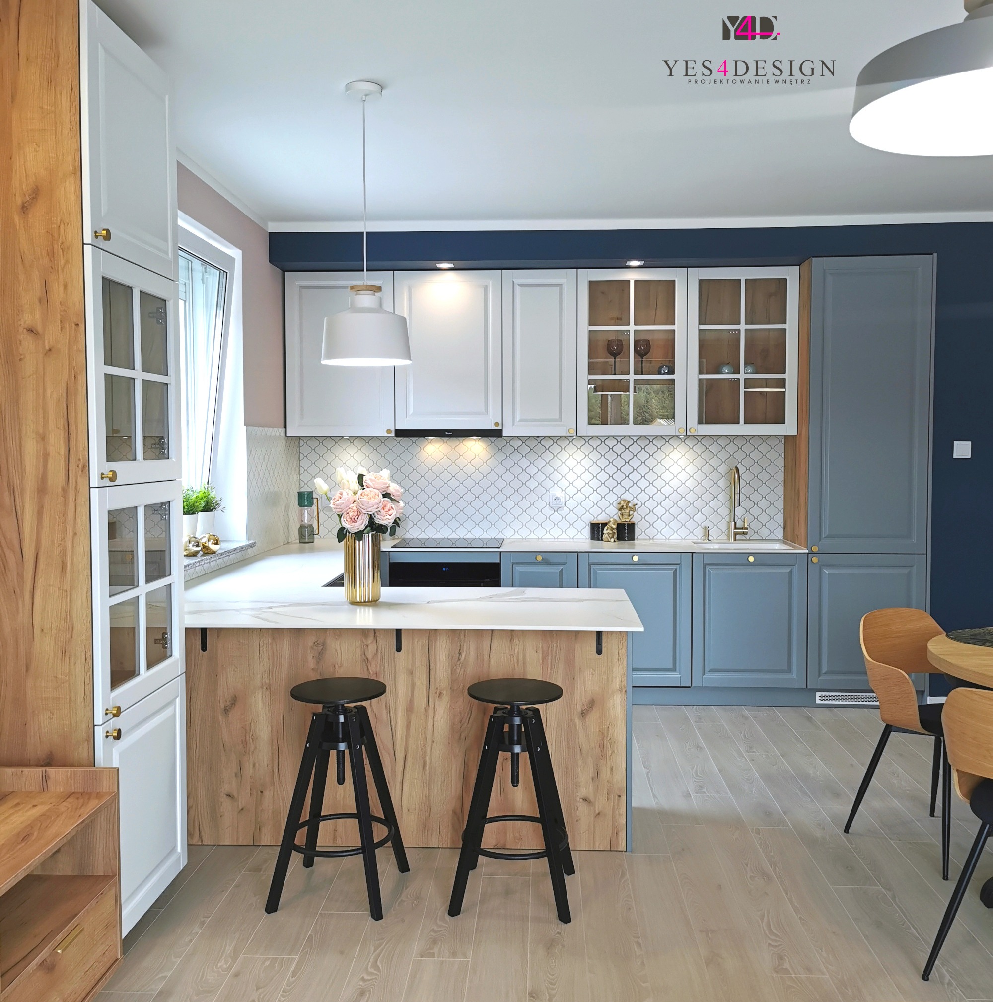 Jasna kuchnia z wyspą, drewnianymi akcentami i czarnymi hokerami, złote detale armatury i uchwytów, kwiaty w złotym wazonie na blacie, aranżacja w stylu nowoczesnym.