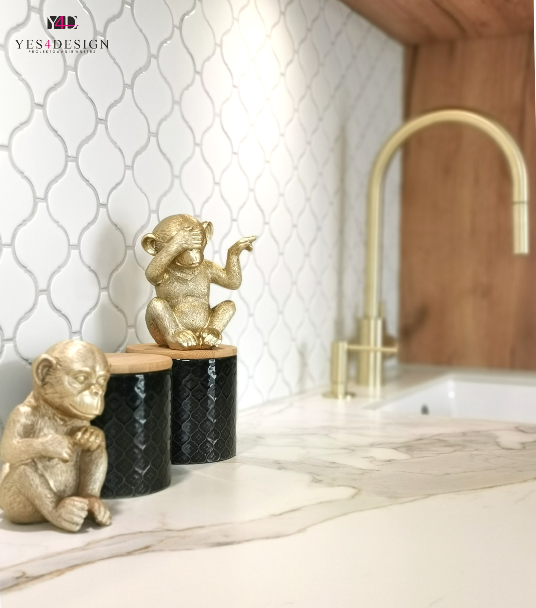 Biała mozaika w kształcie łezki na ścianie nad blatem z marmurowym wzorem; złota bateria umywalkowa i dekoracyjne figurki małp w złotym kolorze na czarnych pojemnikach z bambusowymi pokrywkami.