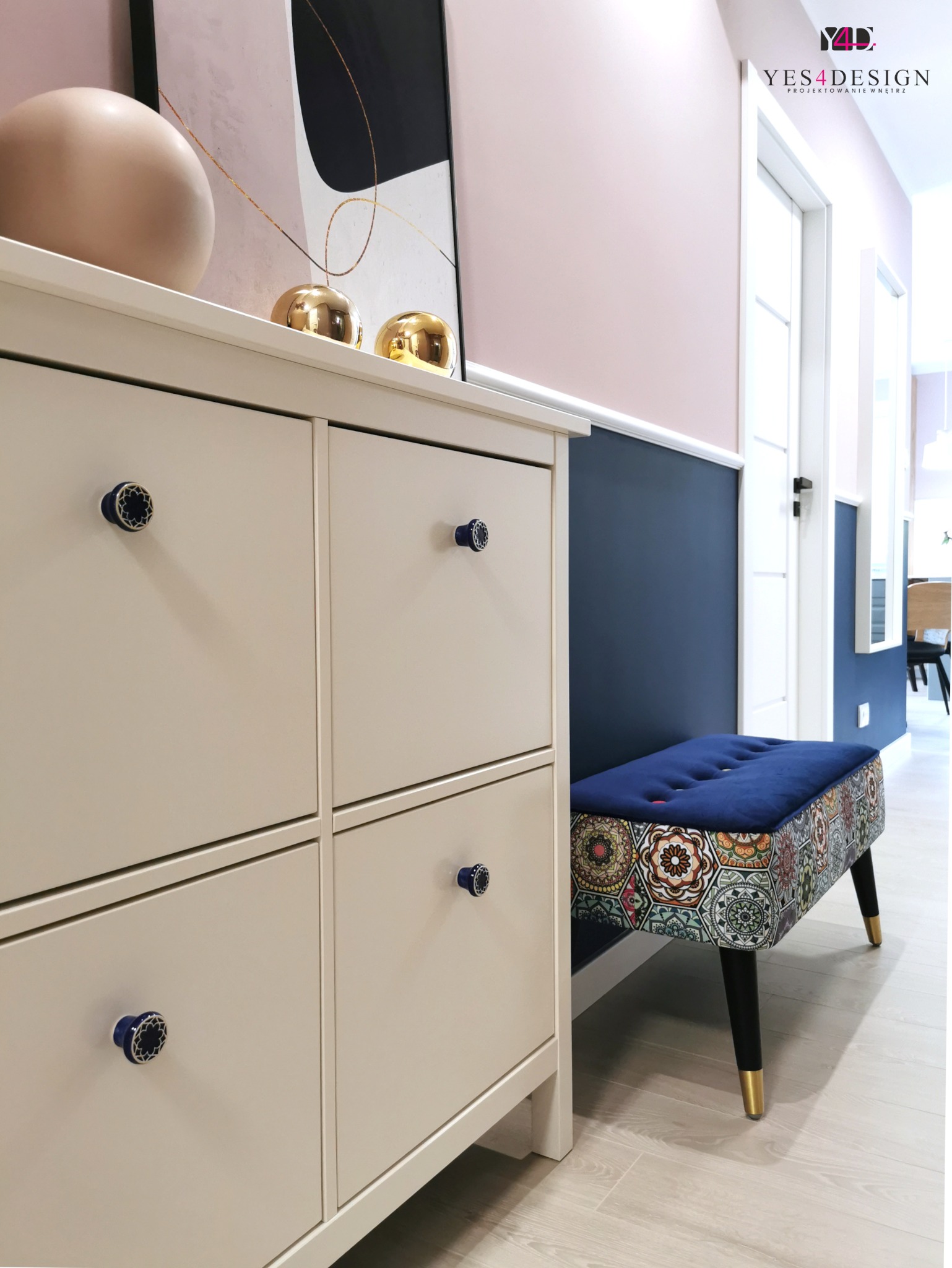 Szary kredens na buty z dekoracyjnymi, granatowymi gałkami, obok tapicerowana pufa z geometrycznym wzorem, na ścianie róż i granat, w tle drzwi i dekoracje.