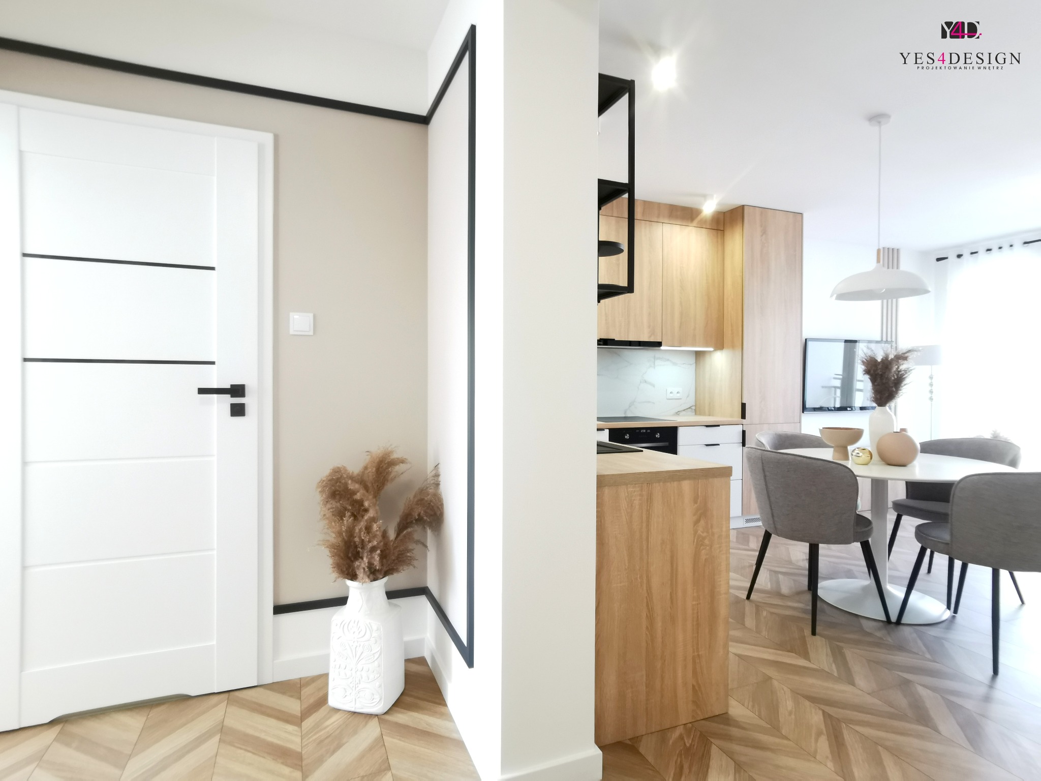 Nowoczesne wnętrze: białe drzwi z czarnymi akcentami, parkiet w jodełkę, dekoracyjny wazon z suszem, otwarta kuchnia z drewnianymi frontami i białą wyspą, okrągły stół z szarymi krzesłami.