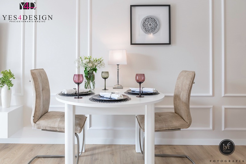 Biały okrągły stół z nakryciem dla dwóch osób, różowe i zielone kieliszki, bukiet białych kwiatów, lampka nocna, dekoracja ścienna w ramce, krzesła z beżową tapicerką w minimalistycznym wnętrzu.