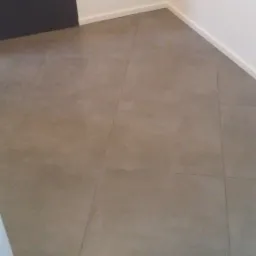 Ułożone kwadratowe panele podłogowe w odcieniach szarości, widoczne fragmenty białej ściany z listwą przypodłogową i ciemne drzwi w rogu.