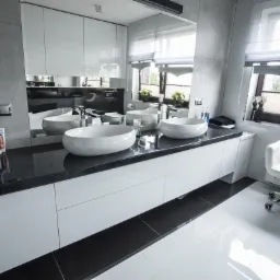 Nowoczesna łazienka z trzema umywalkami nablatowymi na czarnym blacie, białe szafki pod umywalkami, białe płytki podłogowe przechodzące w czarne, białe szafki wiszące z lustrami, białe krzesło...