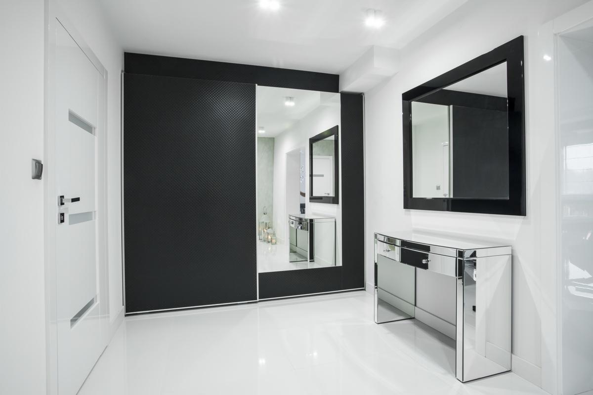 Nowoczesny przedpokój w czerni i bieli z przesuwną szafą z lustrem i konsolą z lustrzanym wykończeniem, oświetlony punktowymi lampami.