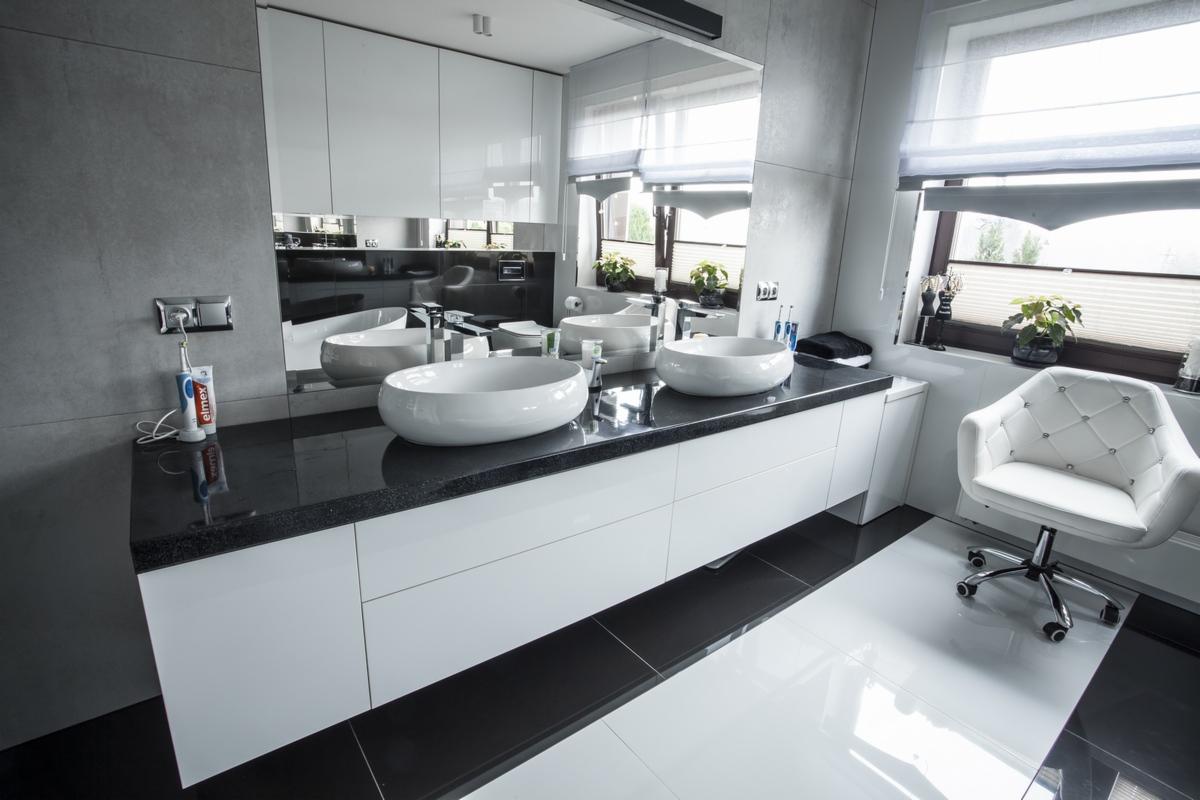 Nowoczesna łazienka z trzema umywalkami nablatowymi na czarnym blacie, białe szafki pod umywalkami, białe płytki podłogowe przechodzące w czarne, białe szafki wiszące z lustrami, białe krzesło...