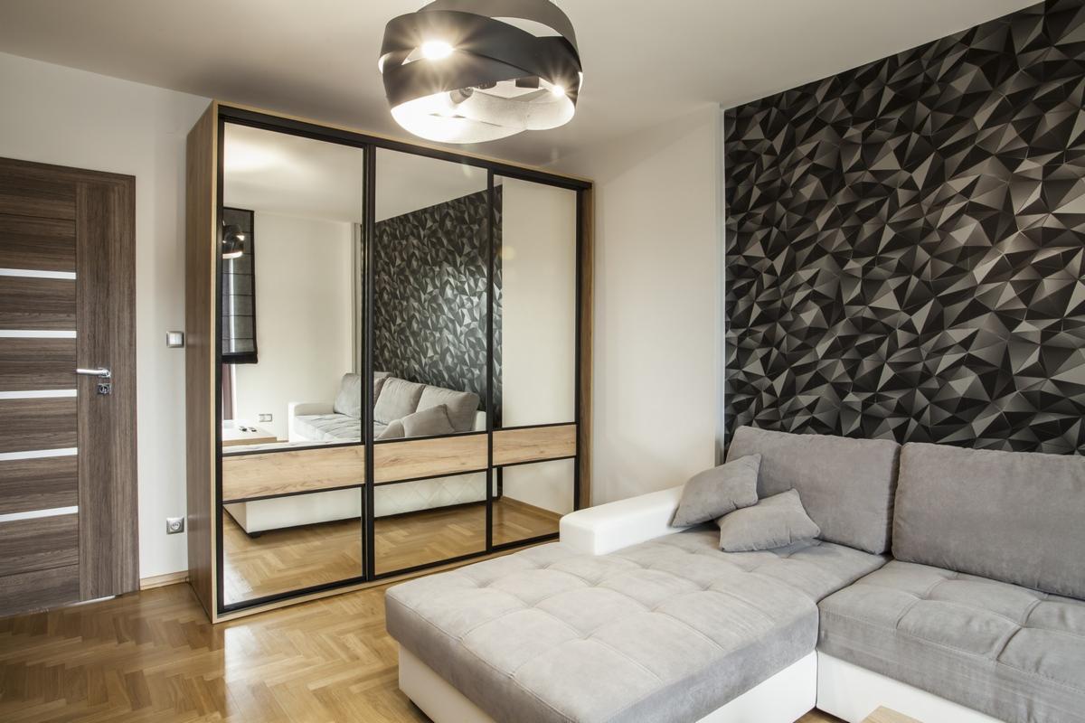 Nowoczesna szafa przesuwna z lustrami w czarnych ramach, z drewnianymi szufladami, odbijająca szarą sofę w salonie z geometryczną tapetą i parkietem w jodełkę.