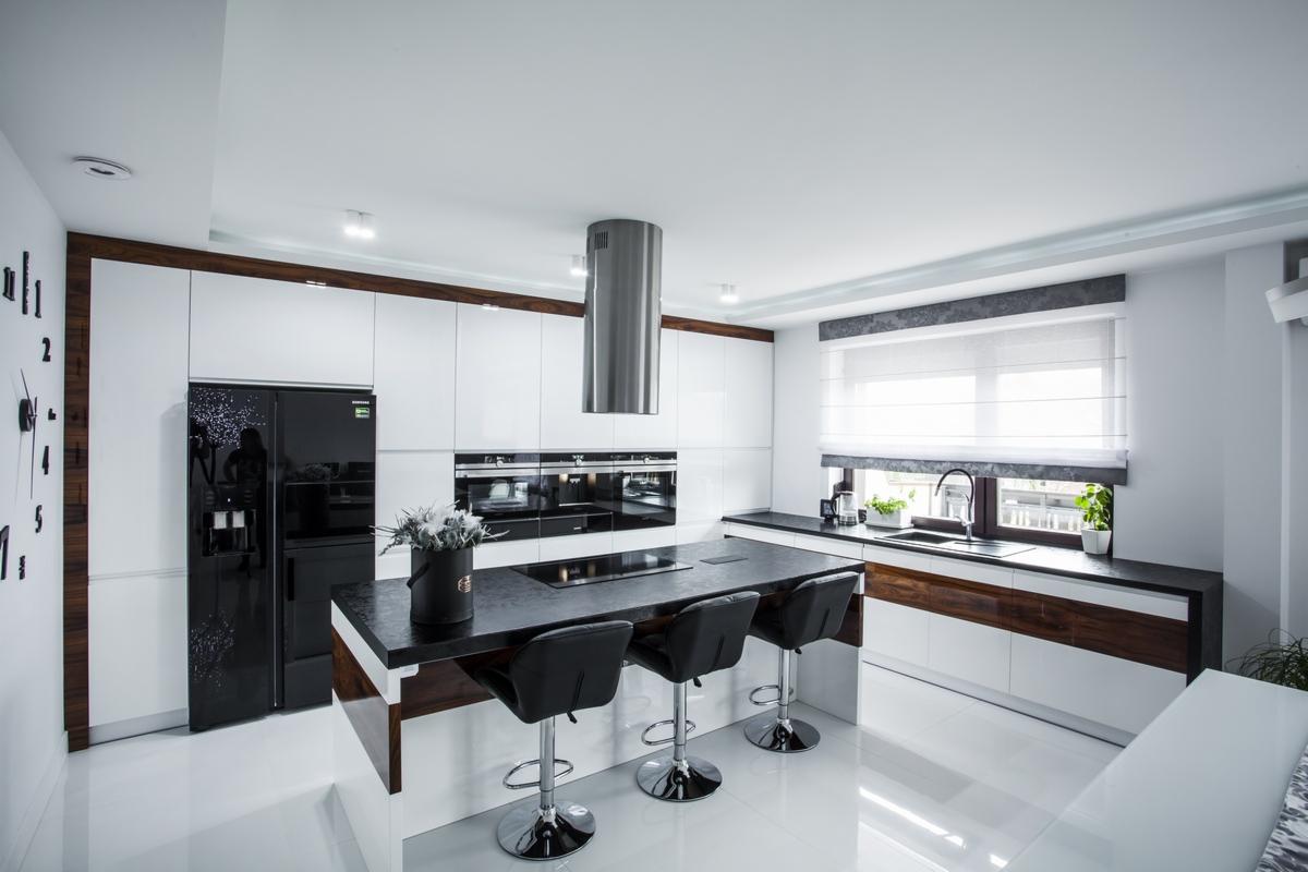 Nowoczesna kuchnia z białymi szafkami, czarnym blatem i wyspą z trzema hokerami, oświetlona naturalnym światłem z okna z roletą rzymską, z dekoracyjnym zegarem na ścianie.