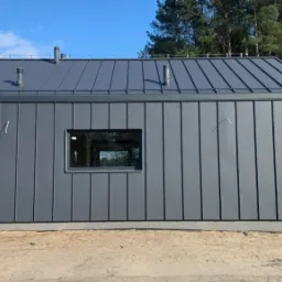 Szary dom z blachy na rąbek stojący, widok z boku, z elementami instalacji wystającymi ze ściany, na tle lasu i niebieskiego nieba.