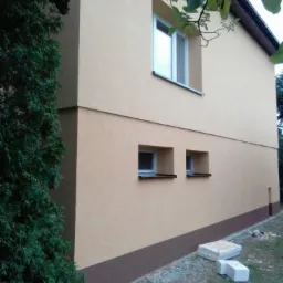 Dwukondygnacyjny budynek z beżową elewacją i brązowym cokołem, widoczne dwa małe okna na parterze i jedno większe na piętrze, obok rosnący krzew i kilka betonowych bloczków na trawie.