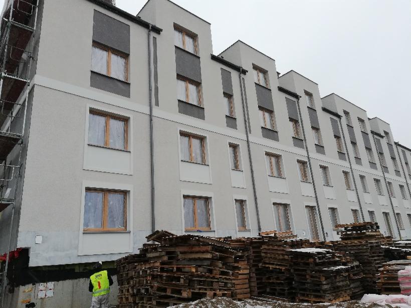 Wielopiętrowy budynek mieszkalny w trakcie prac wykończeniowych elewacji, widoczne rusztowania, palety z materiałami budowlanymi i pracownik w odblaskowej kamizelce.