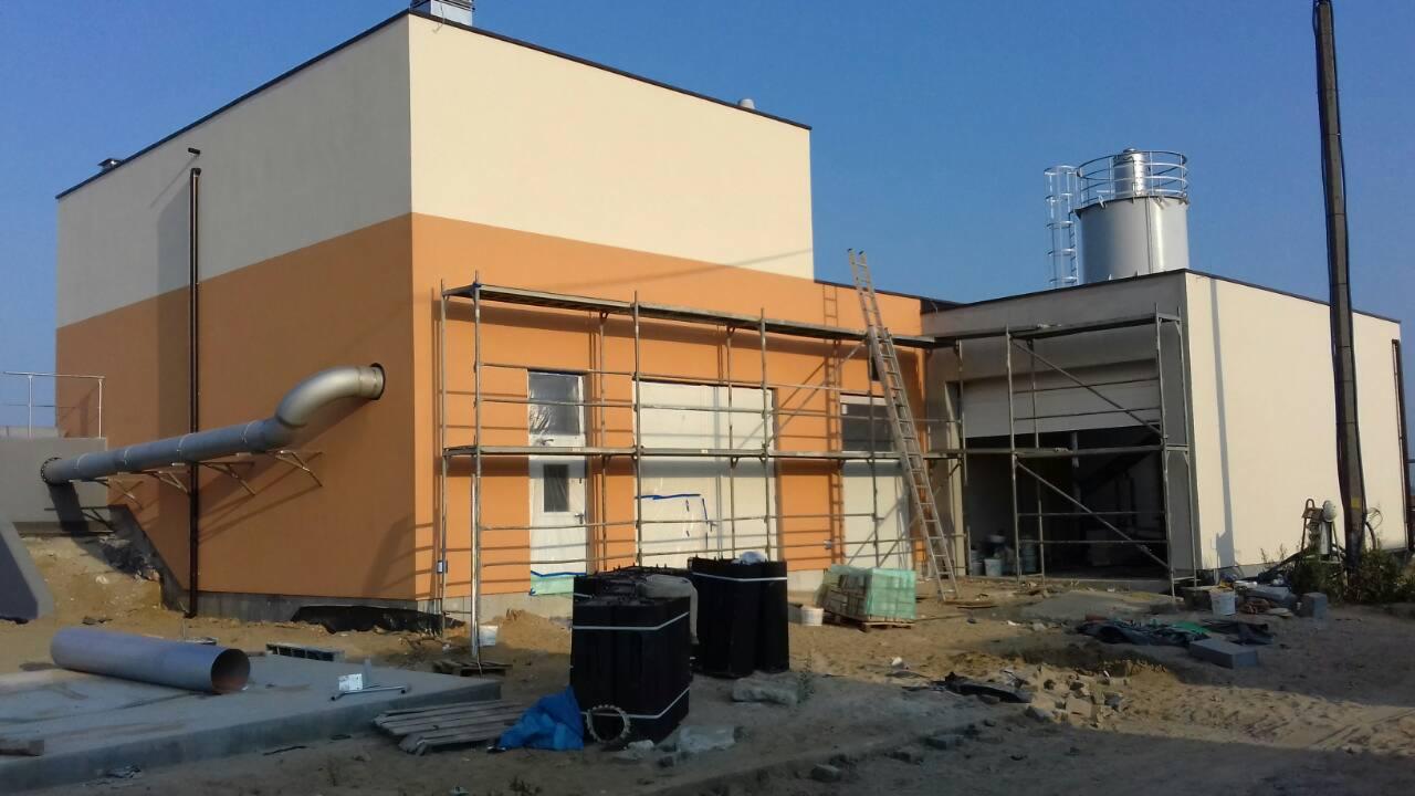 Ocieplona i pomalowana elewacja budynku przemysłowego w trakcie prac wykończeniowych, widoczne rusztowania, rury wentylacyjne i materiały budowlane.
