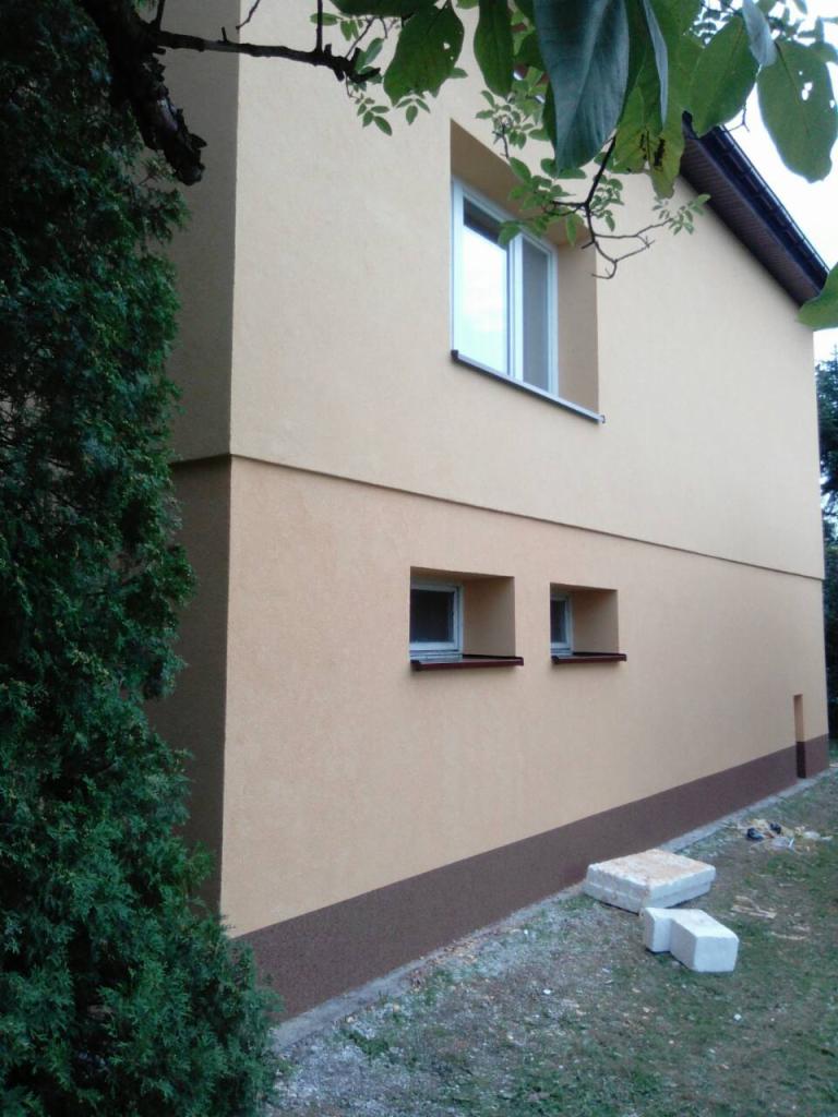 Dwukondygnacyjny budynek z beżową elewacją i brązowym cokołem, widoczne dwa małe okna na parterze i jedno większe na piętrze, obok rosnący krzew i kilka betonowych bloczków na trawie.