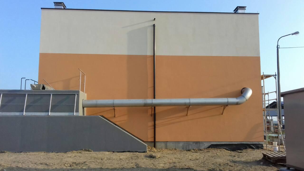 Dwukolorowa elewacja budynku z szarym elementem konstrukcyjnym i metalową rurą wentylacyjną na zewnątrz, widoczna drabina rusztowania i lampa uliczna.