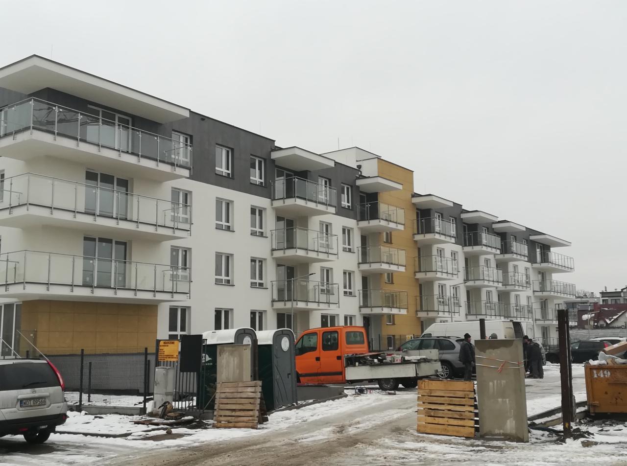 Nowo wybudowany budynek wielorodzinny z balkonami, widok z poziomu ulicy, z tymczasowymi toaletami i pomarańczowym samochodem dostawczym na pierwszym planie, teren budowy w zimowych warunkach.