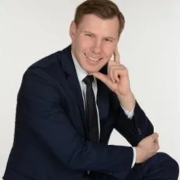 Uśmiechnięty mężczyzna w ciemnym garniturze i krawacie, opierający palec o policzek, na jasnym tle.