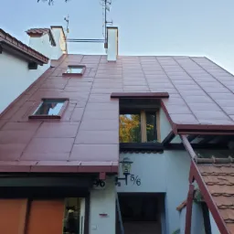 Czerwony dach z blachy z oknami dachowymi i kominami, widok z dołu na budynek z numerem 5/6, widoczne farby i drabina.