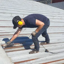 Pracownik w słuchawkach ochronnych szlifuje metalowy dach falisty szlifierką kątową, przygotowując go do malowania lub naprawy, widoczne ślady korozji.