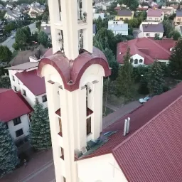 Prace wysokościowe przy wieży kościelnej w Rzeszowie, widok z góry na robotników zabezpieczonych linami. Fragment dachu kościoła z blachy trapezowej w kolorze czerwonym.