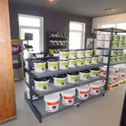 Ekspozycja puszek z farbą na metalowych regałach w składzie budowlanym, widoczne różne kolory i pojemności, w tle okna i inne regały z produktami.