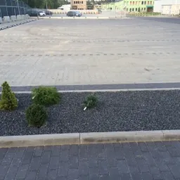 Duży parking wyłożony jasnoszarą kostką brukową, oddzielony od strefy zieleni obrzeżem z ciemnej kostki. Wzdłuż parkingu rząd małych, ozdobnych iglaków posadzonych w czarnym żwirze, ograniczonych...