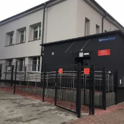 Dwukondygnacyjny budynek po remoncie z szarą elewacją i czarnym dobudowanym segmentem, z rampą dla niepełnosprawnych zabezpieczoną czarnym, metalowym ogrodzeniem i kostką brukową. Tabliczka z nazwą...