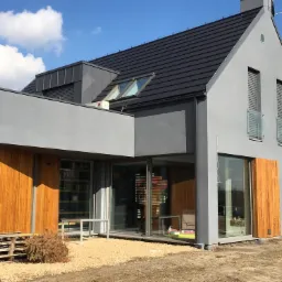 Nowoczesny dom murowany z szarą elewacją, drewnianymi panelami, czarnym dachem i oknami dachowymi, otoczony żwirowym podłożem i paletą.