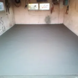 Świeżo wylana, gładka, szara posadzka w pomieszczeniu z oknami i graffiti na ścianach, widoczne ślady po narzędziach w narożnikach.